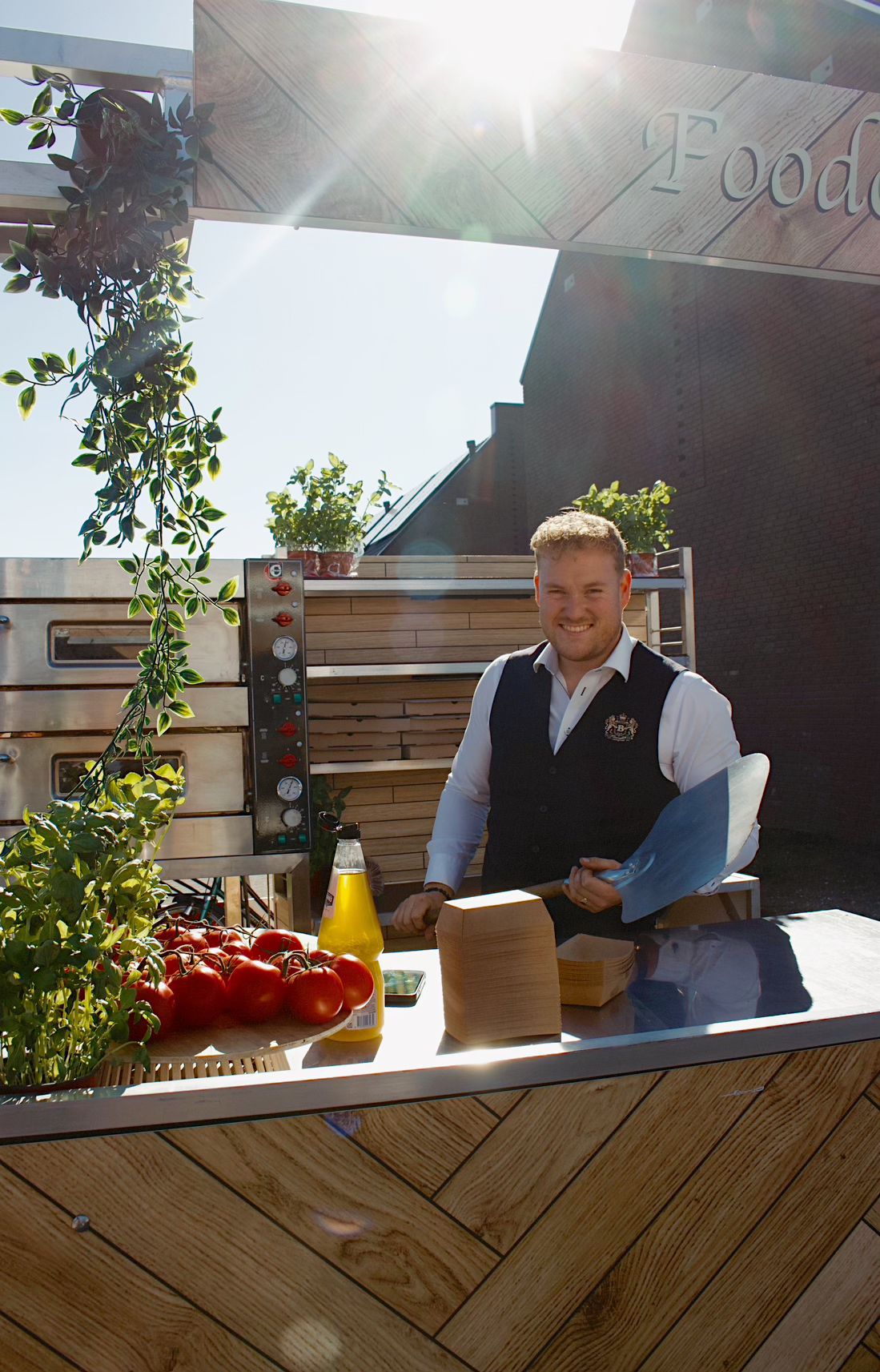 Glimlachende chef bij een pizza-oven met verse tomaten en basilicum.