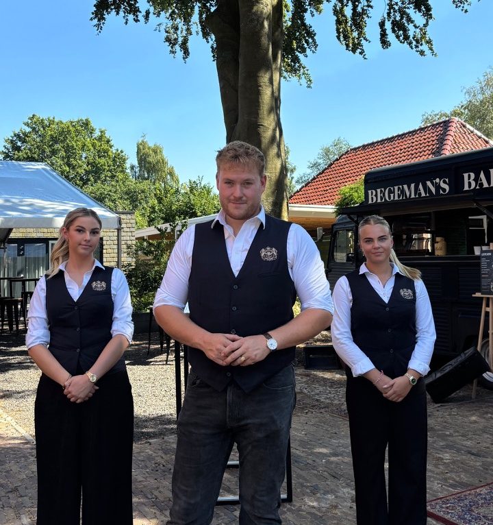 Drie horecamedewerkers poseren buiten onder een grote boom